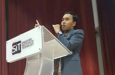 Adjie speaking at a podium of the Singapore Institute of Technology