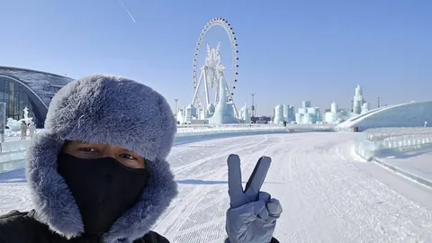 Adjie in a fur hat and face mask, making a peace sign in front of the ice sculptures and the big wheel at the Harbin Ice and Snow World