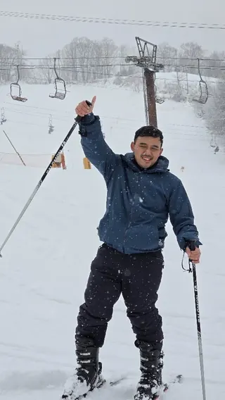 Adjie on skis in falling snow, raising a ski pole with a thumbs up, chairlift behind