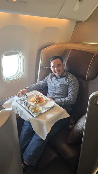 Adjie smiling in a Singapore Airlines First Class suite, with a meal served at the seat by the window